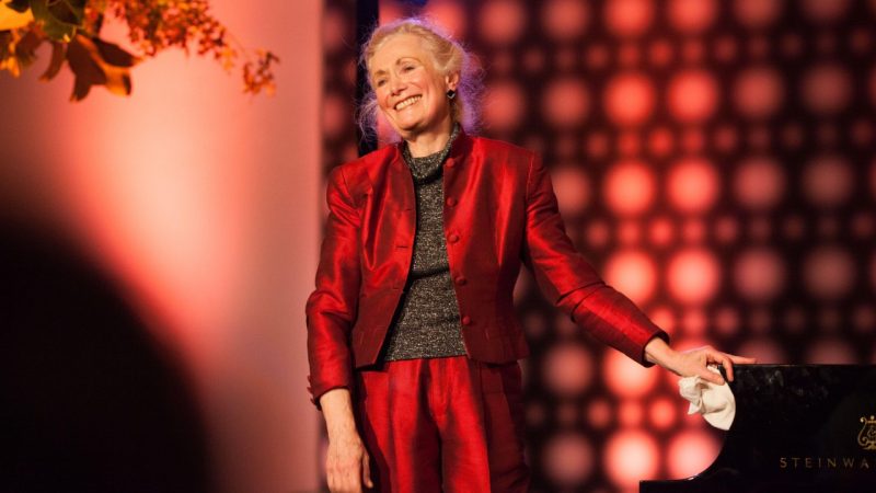Pianist Barbara Nissman, an older white woman with blonde hair pulled into a bun, wears a red silk suit and grey sweater and stands with one hand on a Steinway piano.